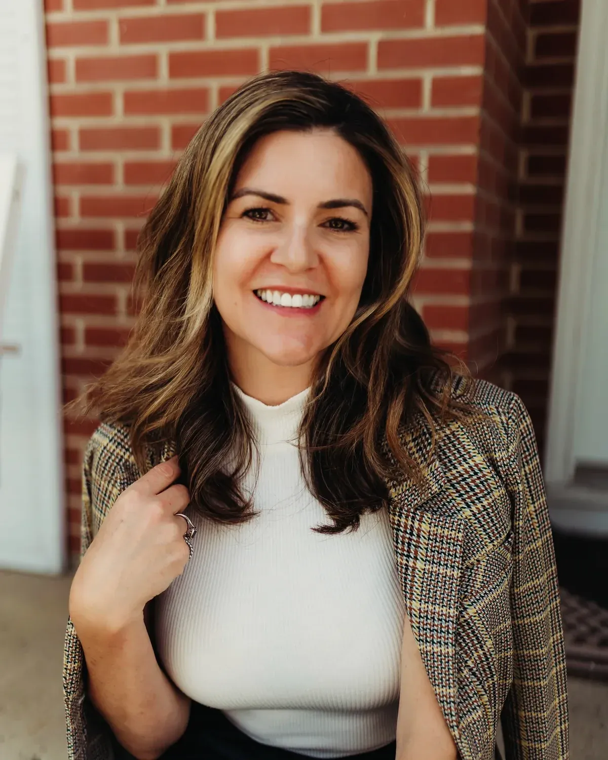 Jess Head smiling in a plaid blazer and white turtleneck against a brick wall